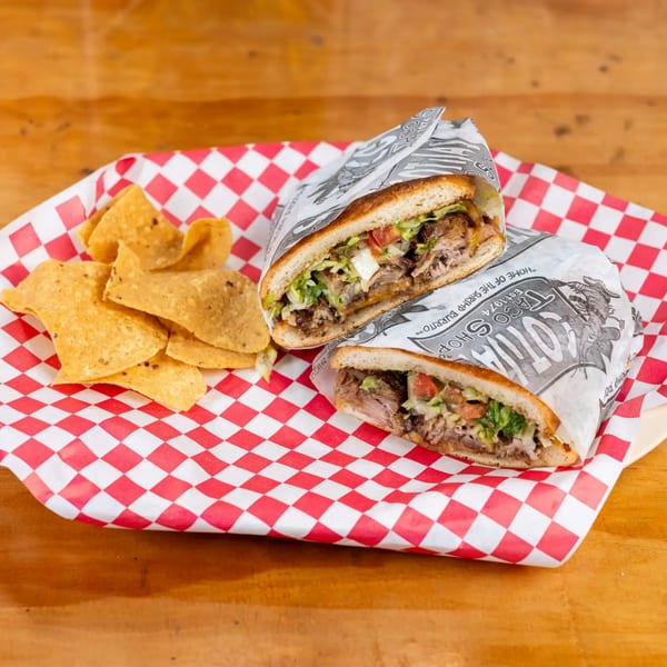 Two halves of a large sandwich wrapped in paper, with tortilla chips on red and white checkered paper.
