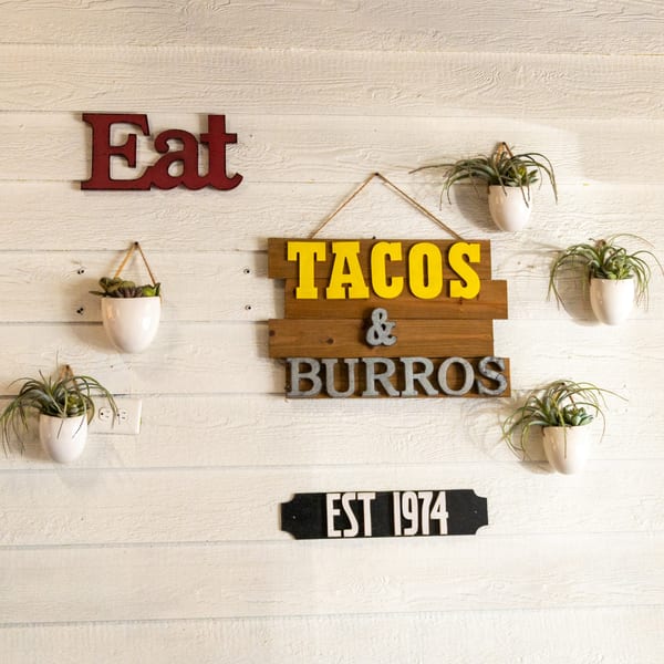 White wooden wall with "Eat Tacos & Burros" signs and potted air plants.