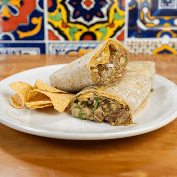 Two burritos with green salsa and tortilla chips on a white plate, against colorful tiled wall.