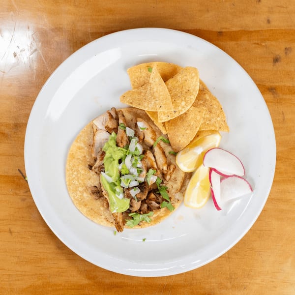 Chicken taco with guacamole, onions, cilantro, tortilla chips, lemon, and radish on a white plate.